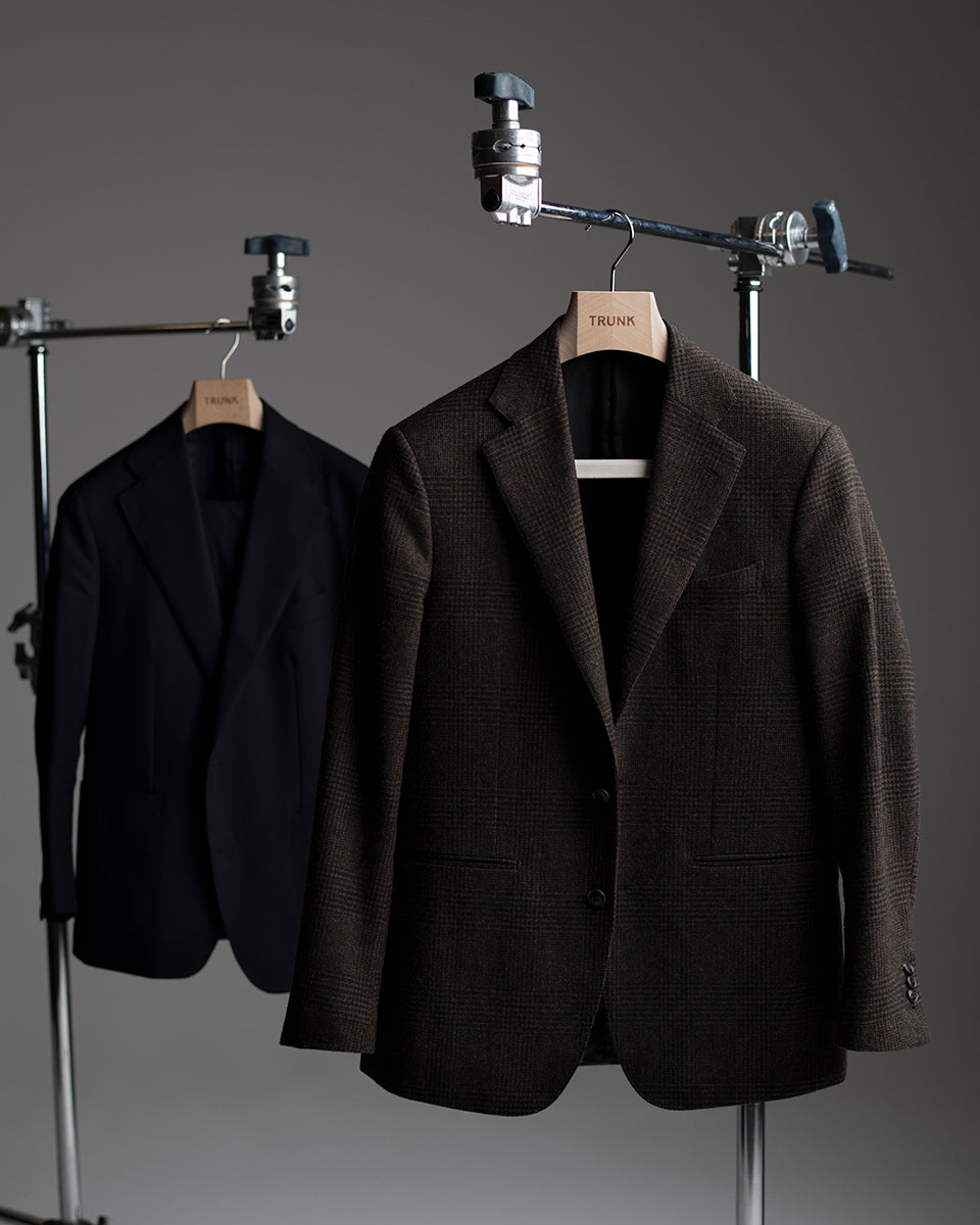 Two suits, one black and one brown, hanging on metal racks against a gray background.