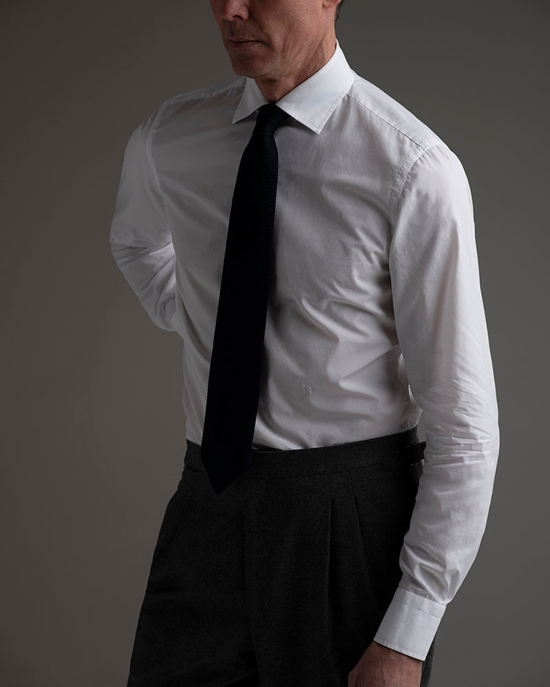 Man wearing a white dress shirt with a black tie and dark pants on a gray background
